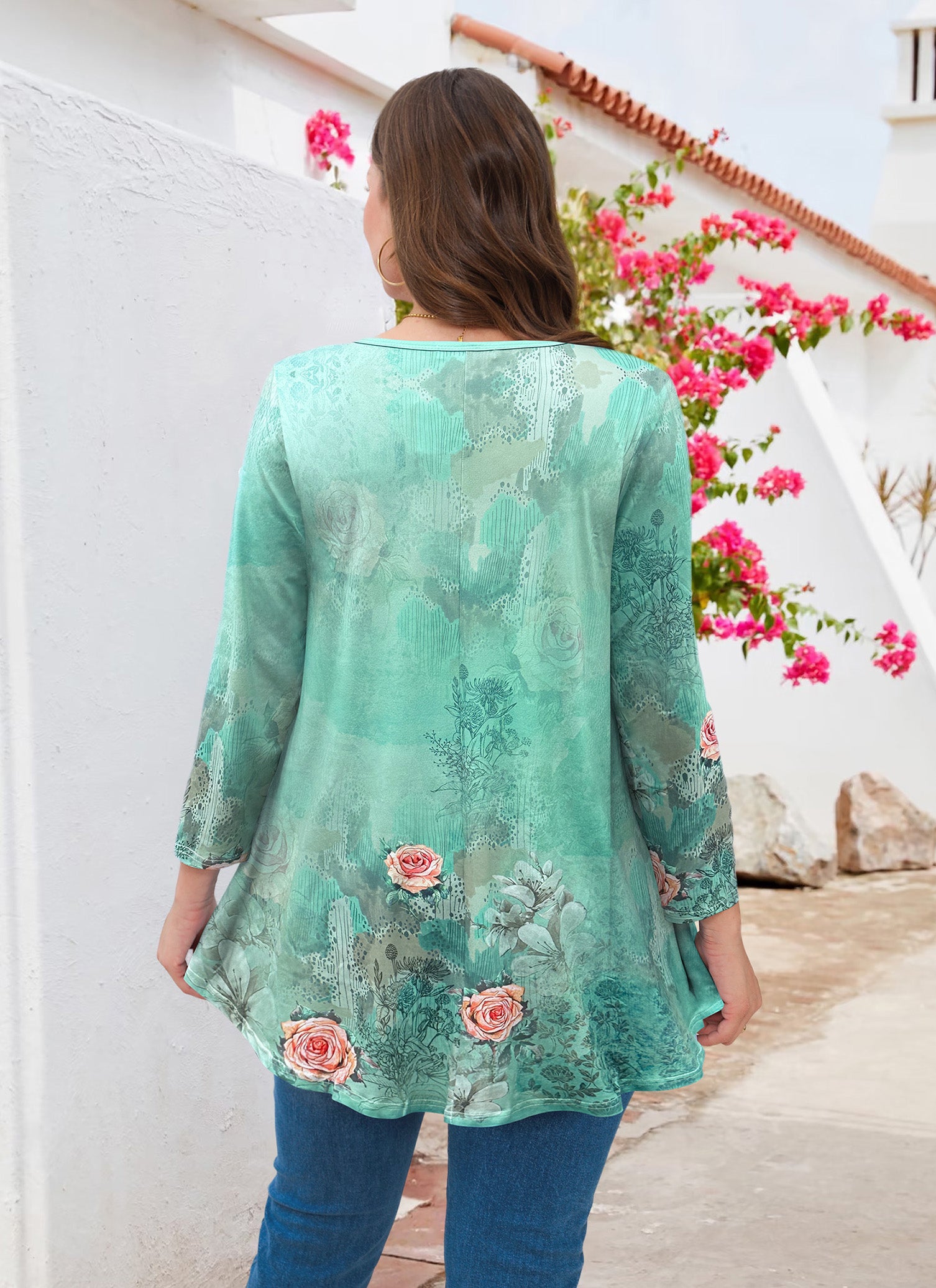 Woman wearing a green floral blouse standing in front of a white wall with pink flowers.